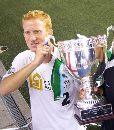 Young Noah Merl on soccer field under stadium lights stands smiling, holding up large trophy for winning Cascadia Cup